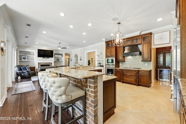 kitchen featuring stainless steel appliances, under cabinet range hood, wine cooler, a brick fireplace, and ornamental molding