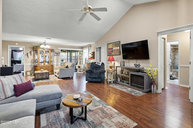 living room with wood finished floors, a textured ceiling, a fireplace with flush hearth, ceiling fan with notable chandelier, and vaulted ceiling