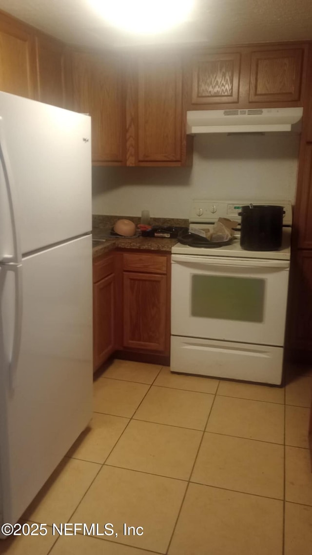kitchen featuring brown cabinets, dark countertops, white appliances, light tile patterned floors, and under cabinet range hood