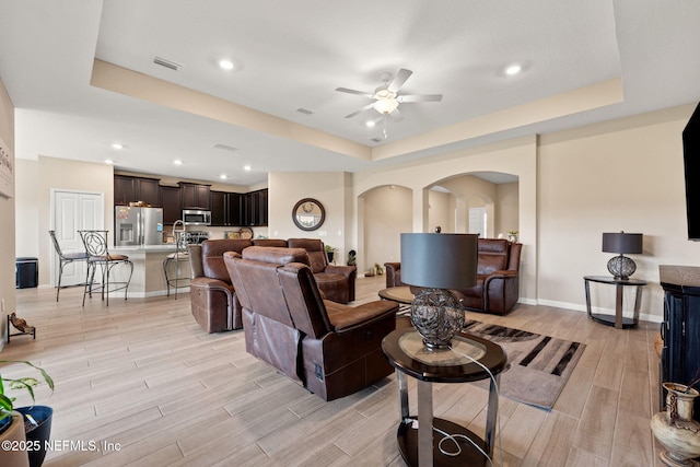 living room with a ceiling fan, arched walkways, baseboards, a tray ceiling, and light wood-style flooring