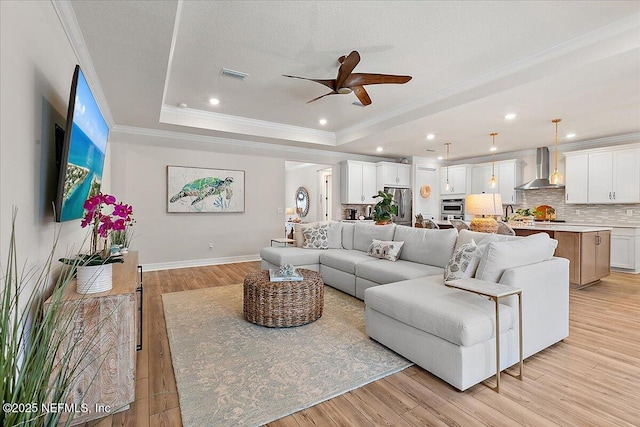 living room with ceiling fan, light wood finished floors, a raised ceiling, visible vents, and ornamental molding