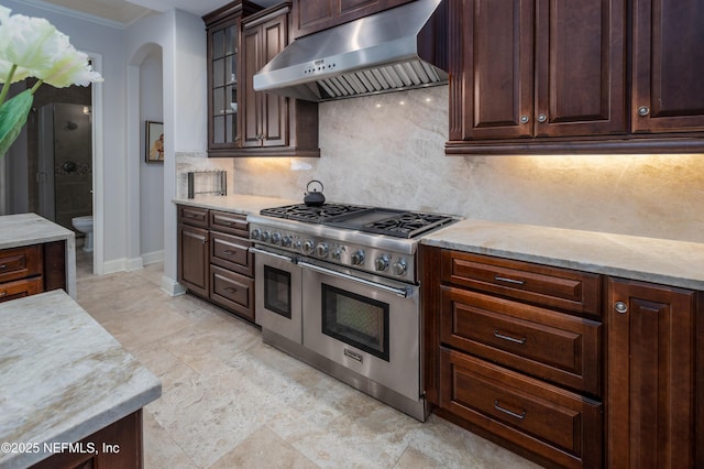 kitchen featuring dark brown cabinets, double oven range, ventilation hood, arched walkways, and tasteful backsplash