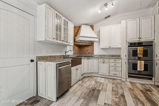 kitchen featuring stainless steel appliances, custom range hood, light wood-style flooring, glass insert cabinets, and a textured ceiling