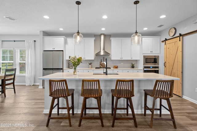 kitchen with wall chimney range hood, stainless steel appliances, baseboards, an island with sink, and light countertops