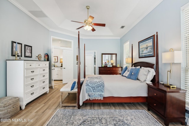 bedroom featuring wood finished floors, ornamental molding, a raised ceiling, ensuite bath, and a ceiling fan