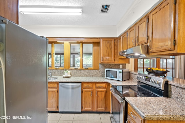 kitchen featuring stainless steel appliances, ornamental molding, brown cabinets, light tile patterned flooring, and under cabinet range hood
