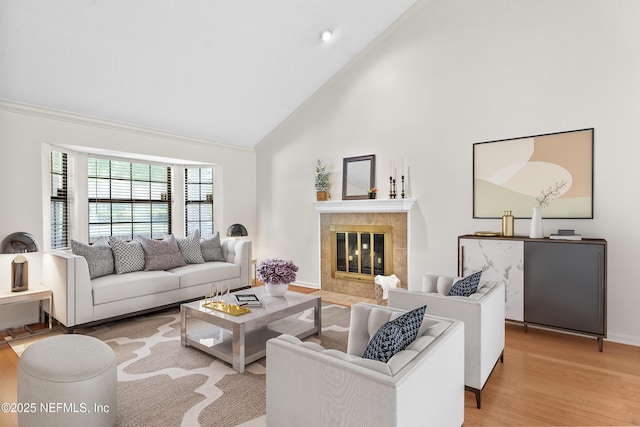 living area featuring high vaulted ceiling, light wood finished floors, a tiled fireplace, crown molding, and baseboards