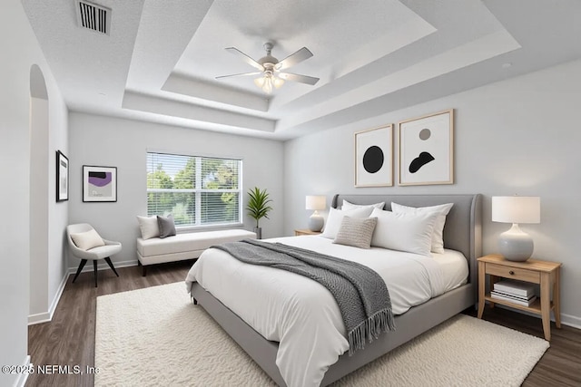 bedroom featuring a raised ceiling, arched walkways, dark wood-style floors, baseboards, and ceiling fan