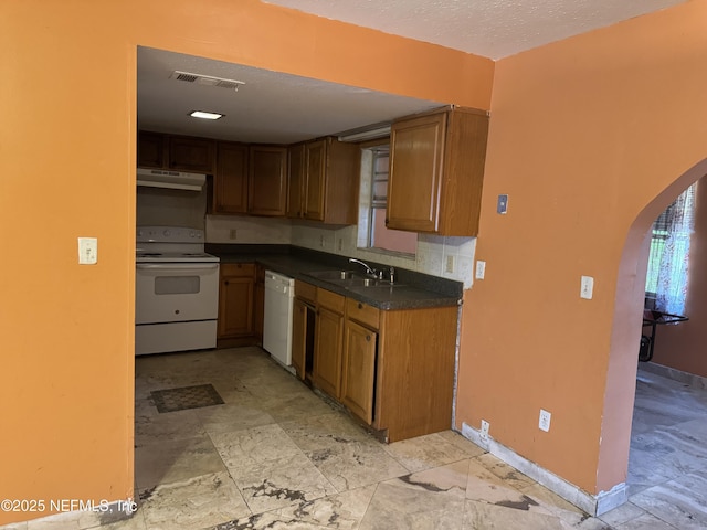 kitchen with white appliances, under cabinet range hood, a sink, arched walkways, and dark countertops