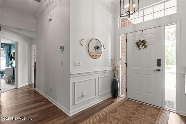 entryway with dark wood-type flooring, a chandelier, ornamental molding, a towering ceiling, and a wainscoted wall