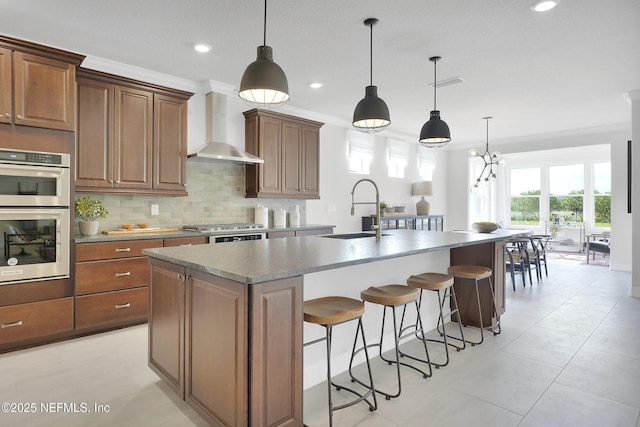 kitchen featuring a sink, wall chimney exhaust hood, stainless steel appliances, a kitchen island with sink, and tasteful backsplash