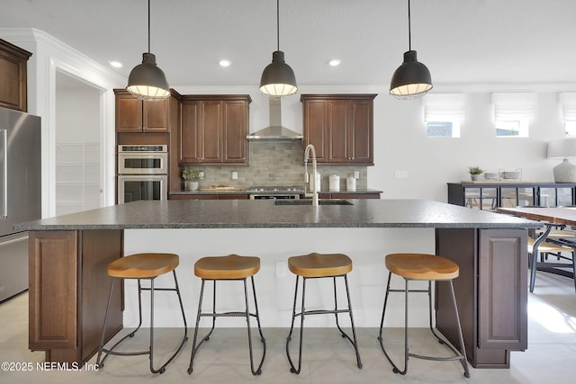 kitchen featuring a sink, stainless steel appliances, dark countertops, wall chimney exhaust hood, and an island with sink