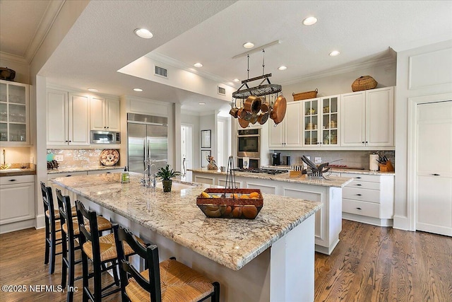 kitchen with a kitchen island with sink, built in appliances, ornamental molding, dark wood finished floors, and tasteful backsplash
