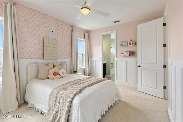 bedroom with a wainscoted wall, light colored carpet, a ceiling fan, connected bathroom, and a decorative wall