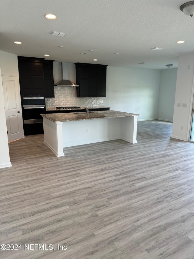 kitchen with wall chimney range hood, open floor plan, light wood-style flooring, a center island with sink, and backsplash