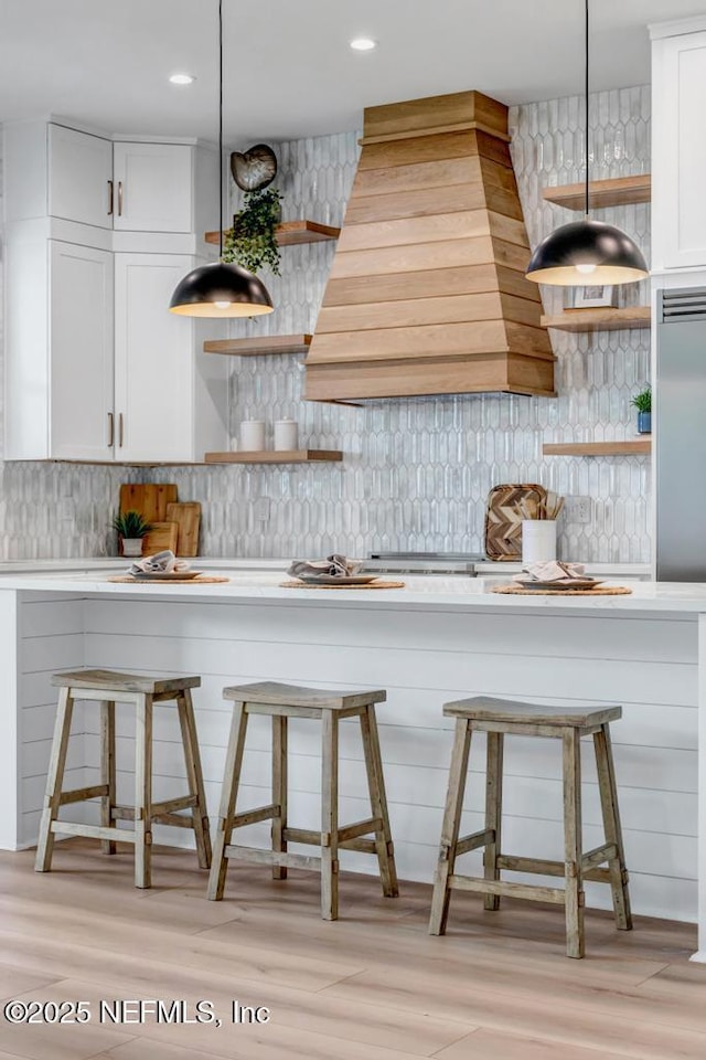 kitchen featuring open shelves, custom range hood, decorative backsplash, a kitchen breakfast bar, and light wood-style floors