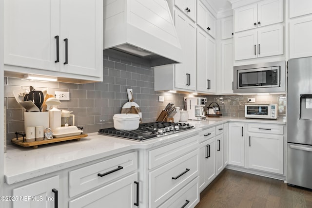 kitchen with appliances with stainless steel finishes, custom range hood, tasteful backsplash, dark wood-type flooring, and white cabinets