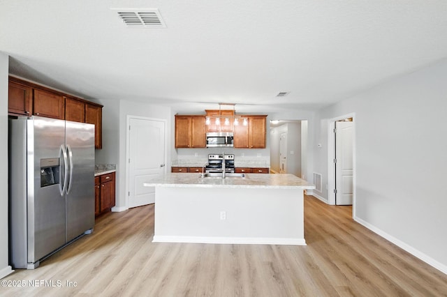 kitchen with appliances with stainless steel finishes, light wood-type flooring, an island with sink, brown cabinets, and baseboards