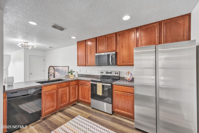 kitchen with appliances with stainless steel finishes, a sink, a textured ceiling, wood finished floors, and dark countertops
