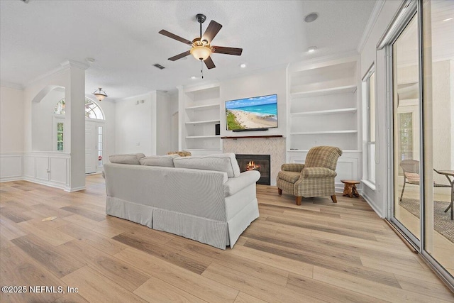 living room featuring ornamental molding, built in features, a glass covered fireplace, a ceiling fan, and light wood-style flooring
