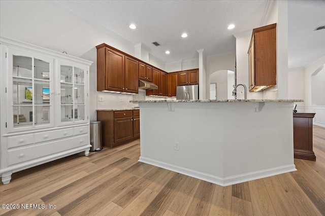 kitchen with stainless steel fridge, under cabinet range hood, crown molding, light stone countertops, and light wood finished floors