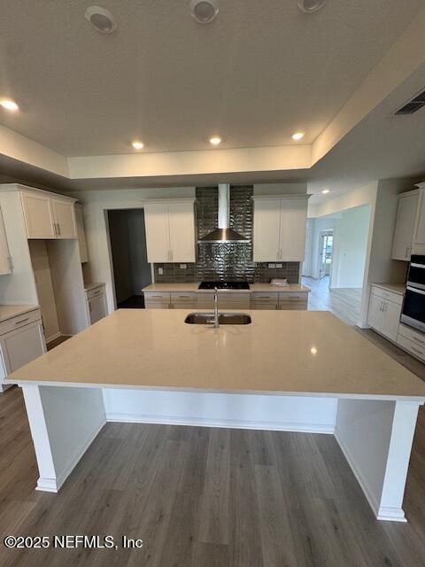 kitchen with wall chimney exhaust hood, a sink, a tray ceiling, a center island with sink, and dark wood finished floors