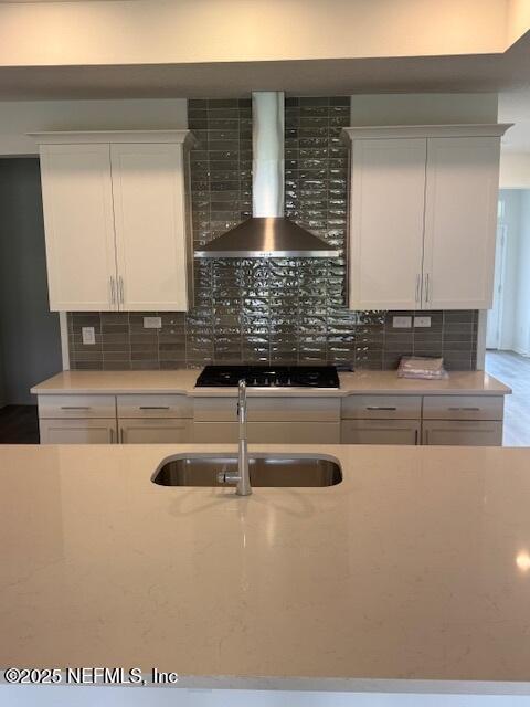 kitchen featuring wall chimney range hood, a sink, decorative backsplash, and white cabinetry