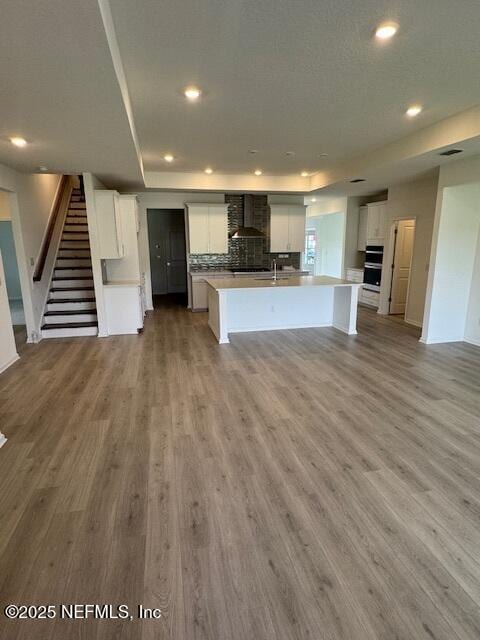 kitchen with wall chimney range hood, light wood-style floors, recessed lighting, an island with sink, and white cabinets