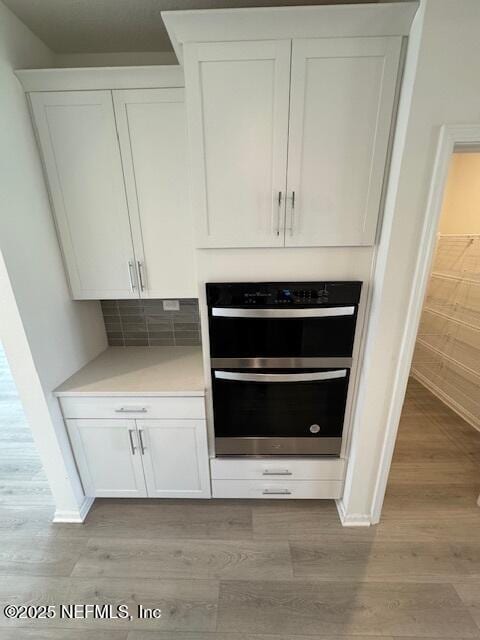 detailed view of double oven, light wood-style flooring, white cabinets, light countertops, and tasteful backsplash