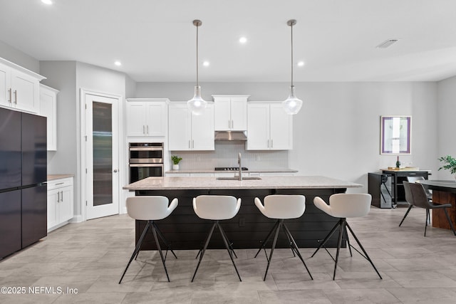 kitchen with freestanding refrigerator, double oven, decorative backsplash, a center island with sink, and under cabinet range hood