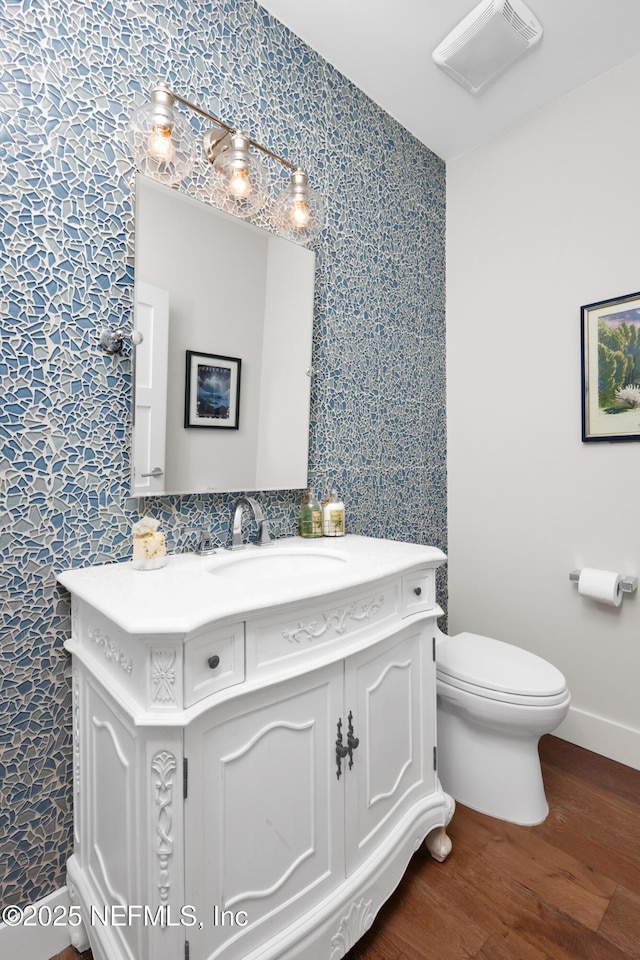 half bathroom featuring wood finished floors, wallpapered walls, toilet, baseboards, and vanity