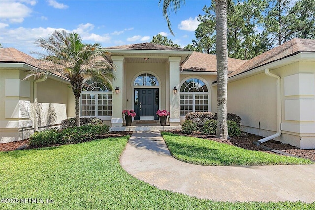 doorway to property featuring stucco siding, a yard, and roof with shingles