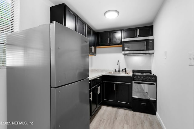kitchen featuring appliances with stainless steel finishes, a sink, dark cabinetry, light wood-type flooring, and light countertops