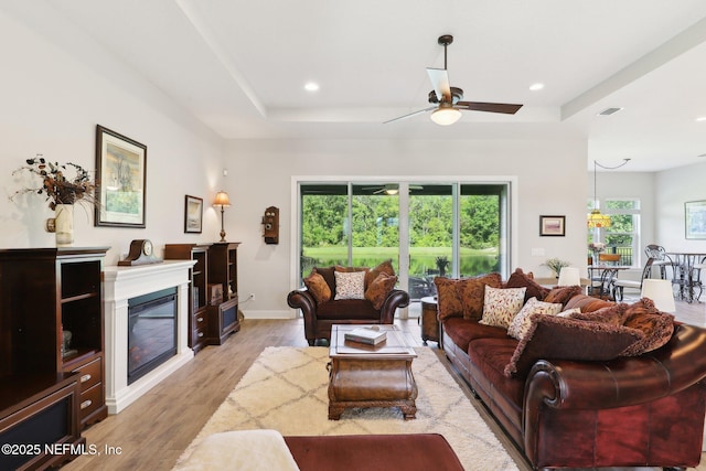 living room featuring a raised ceiling, ceiling fan, light wood finished floors, recessed lighting, and a glass covered fireplace
