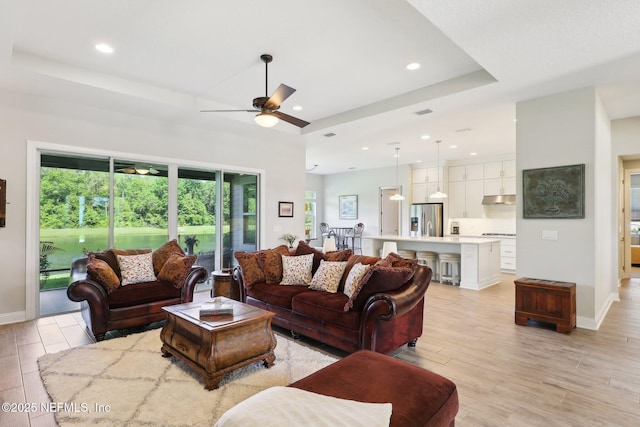 living area with a ceiling fan, light wood-style flooring, a raised ceiling, recessed lighting, and baseboards