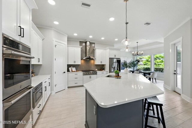 kitchen featuring wall chimney exhaust hood, appliances with stainless steel finishes, a sink, backsplash, and a large island with sink