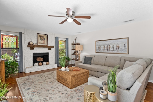 living room with ceiling fan, wood finished floors, a stone fireplace, and a textured ceiling