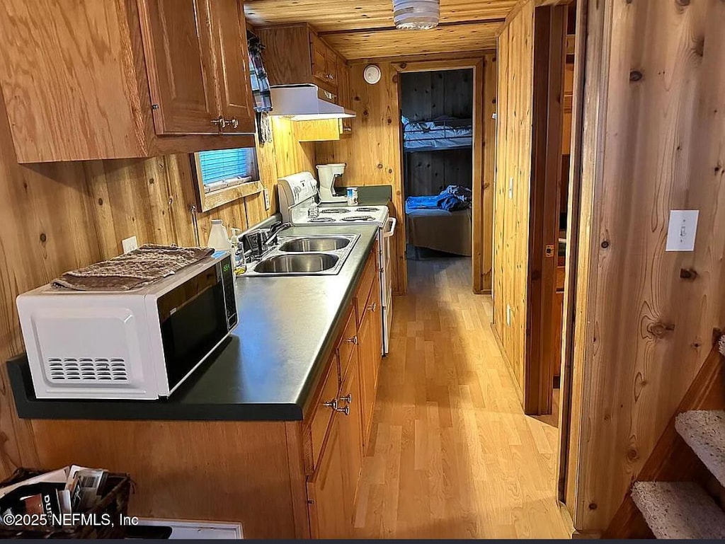 kitchen featuring white microwave, a sink, under cabinet range hood, wooden walls, and light wood-style floors