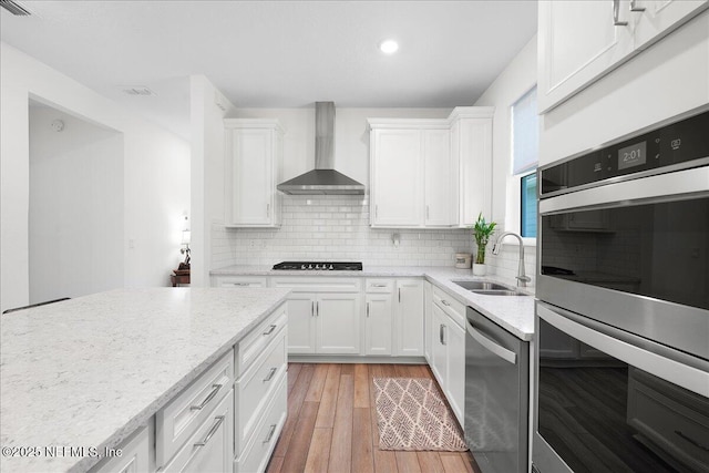 kitchen with a sink, appliances with stainless steel finishes, wall chimney exhaust hood, light wood finished floors, and white cabinetry