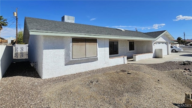 rear view of house with a garage, roof with shingles, fence, and stucco siding