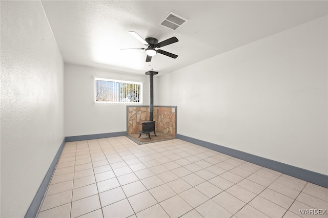 empty room featuring ceiling fan, light tile patterned floors, visible vents, baseboards, and a wood stove