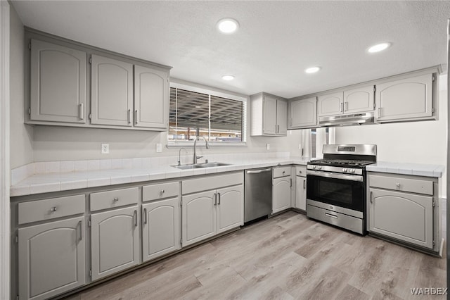 kitchen featuring light wood-style floors, tile countertops, stainless steel appliances, under cabinet range hood, and a sink