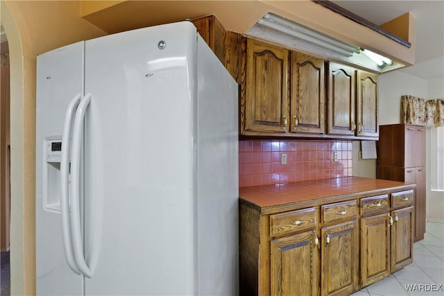 kitchen with light tile patterned floors, tile counters, white fridge with ice dispenser, tasteful backsplash, and brown cabinetry