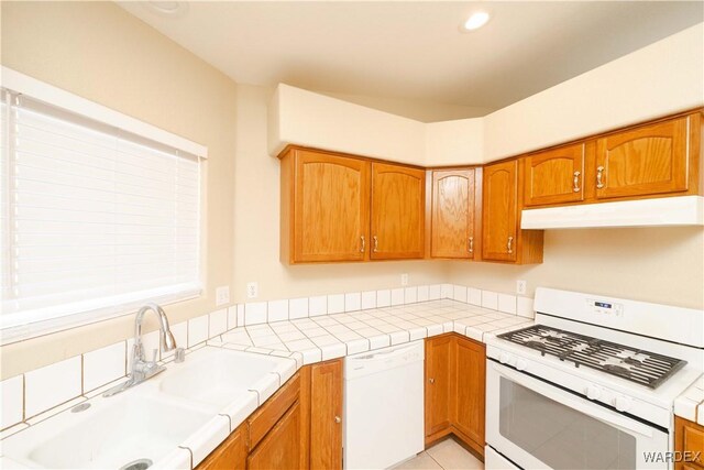 kitchen with white appliances, tile countertops, brown cabinets, under cabinet range hood, and a sink