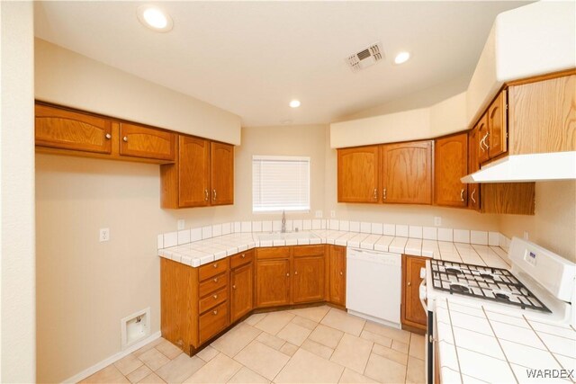 kitchen with under cabinet range hood, white appliances, a sink, tile counters, and brown cabinetry