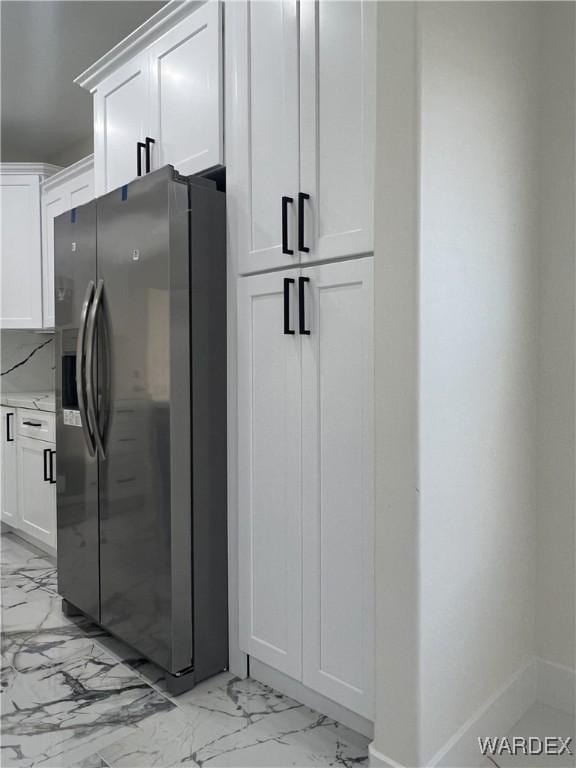 kitchen with light stone counters, marble finish floor, stainless steel fridge with ice dispenser, white cabinets, and baseboards