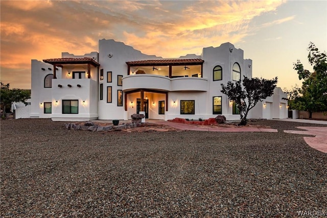 back of property at dusk with driveway, a balcony, ceiling fan, a tiled roof, and stucco siding