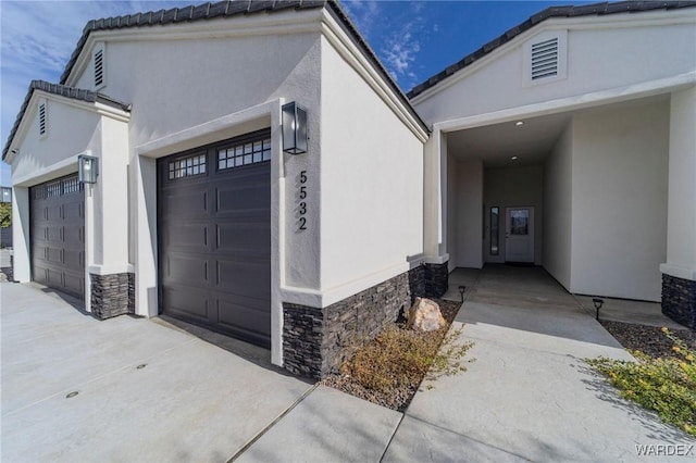 view of exterior entry with stone siding, concrete driveway, visible vents, and stucco siding