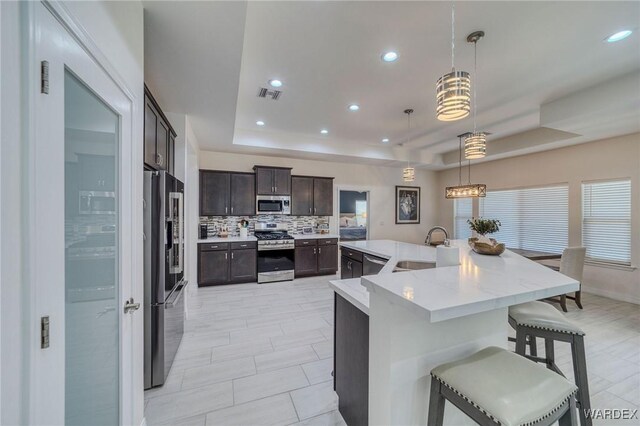 kitchen with a tray ceiling, pendant lighting, a center island with sink, appliances with stainless steel finishes, and a sink