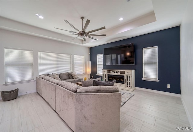 living room featuring baseboards, a raised ceiling, a ceiling fan, a glass covered fireplace, and a wealth of natural light